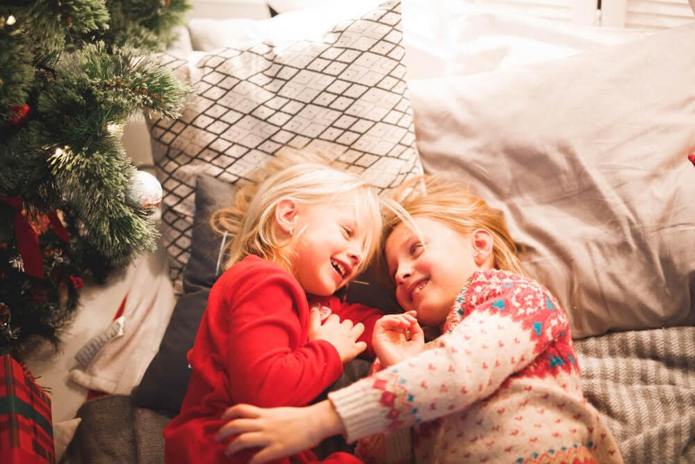 Two Girls Laying Down Hugging Each Other Beside A Christmas Tree
